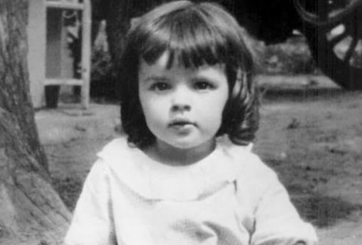 Black-and-white photo of a toddler sitting on the ground outdoors. The child has short dark hair with bangs, wears a light dress with short sleeves, and looks directly at the camera with a calm expression.