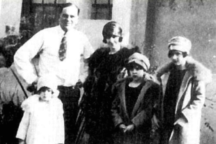 Grainy black-and-white photo of a family from the early 20th century. A man in a white shirt stands beside a woman and three young girls, all wearing cloche hats and winter coats with fur collars.