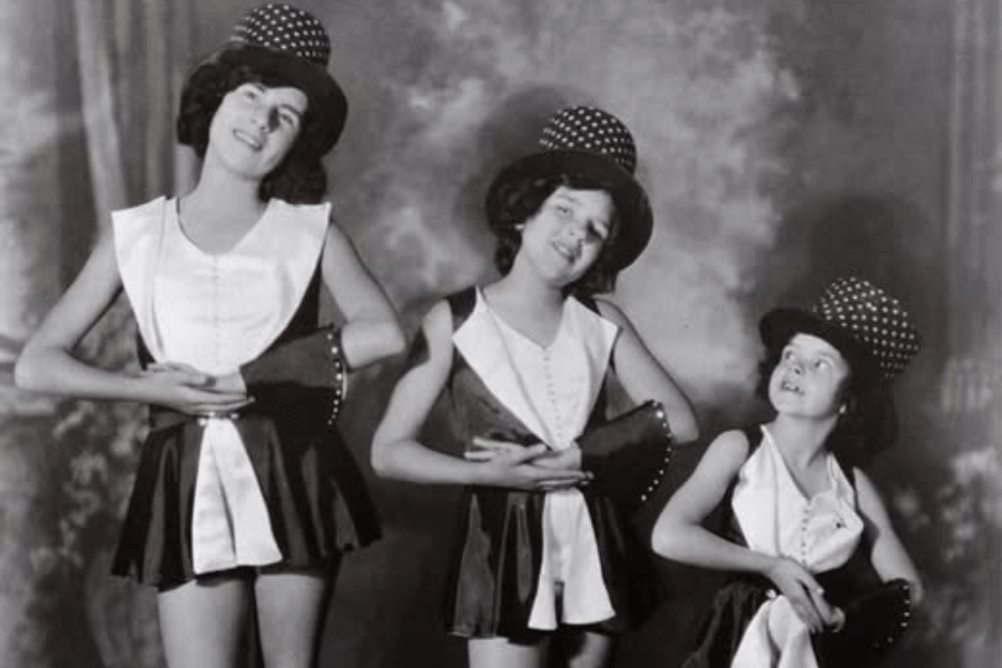 Black-and-white photo of three young girls in matching dance costumes and polka-dot top hats. They pose mid-performance with tilted heads, arms bent in unison, and wide grins, suggesting a theatrical dance number.