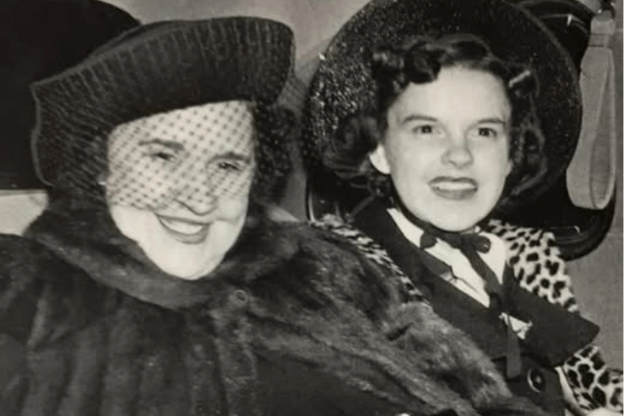 Black-and-white photo of two women sitting in the back of a car. The woman on the left wears a fur coat and a netted hat, while the younger woman on the right smiles brightly in a leopard-print coat and wide-brimmed hat.