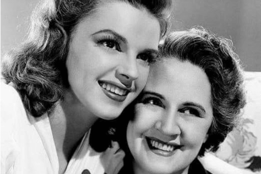 Black-and-white portrait of two smiling women embracing closely. The younger woman rests her cheek against the older woman’s head; both wear elegant outfits, with the older woman in pearls and the younger adorned with a large flower brooch.