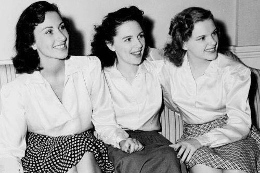 Black-and-white photo of three young women sitting on a striped couch, smiling and holding hands. They wear collared blouses and knee-length skirts, their hairstyles and fashion reflecting the 1940s era.