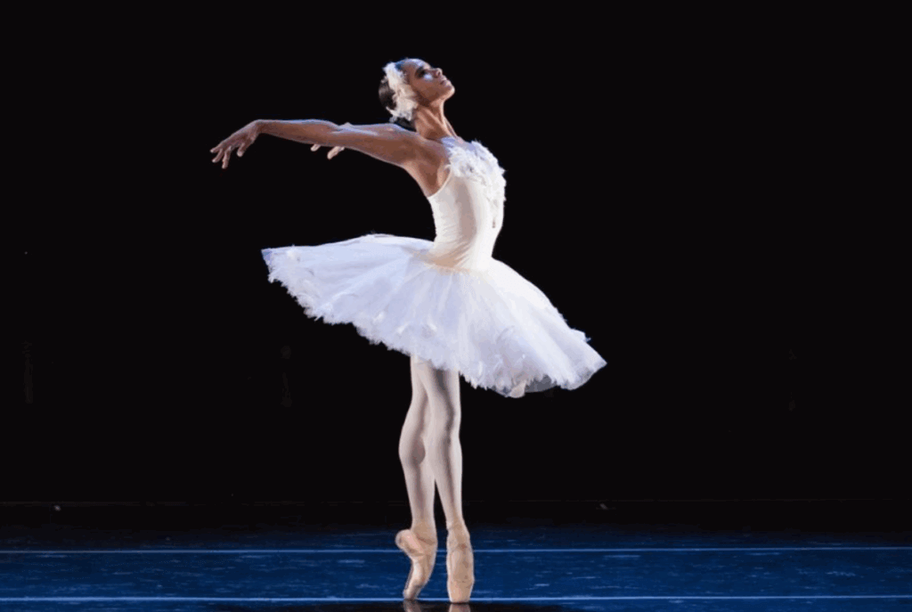 Ballet dancer in a white tutu performs gracefully on stage against a dark background. She is on pointe, arms elegantly extended, conveying elegance.